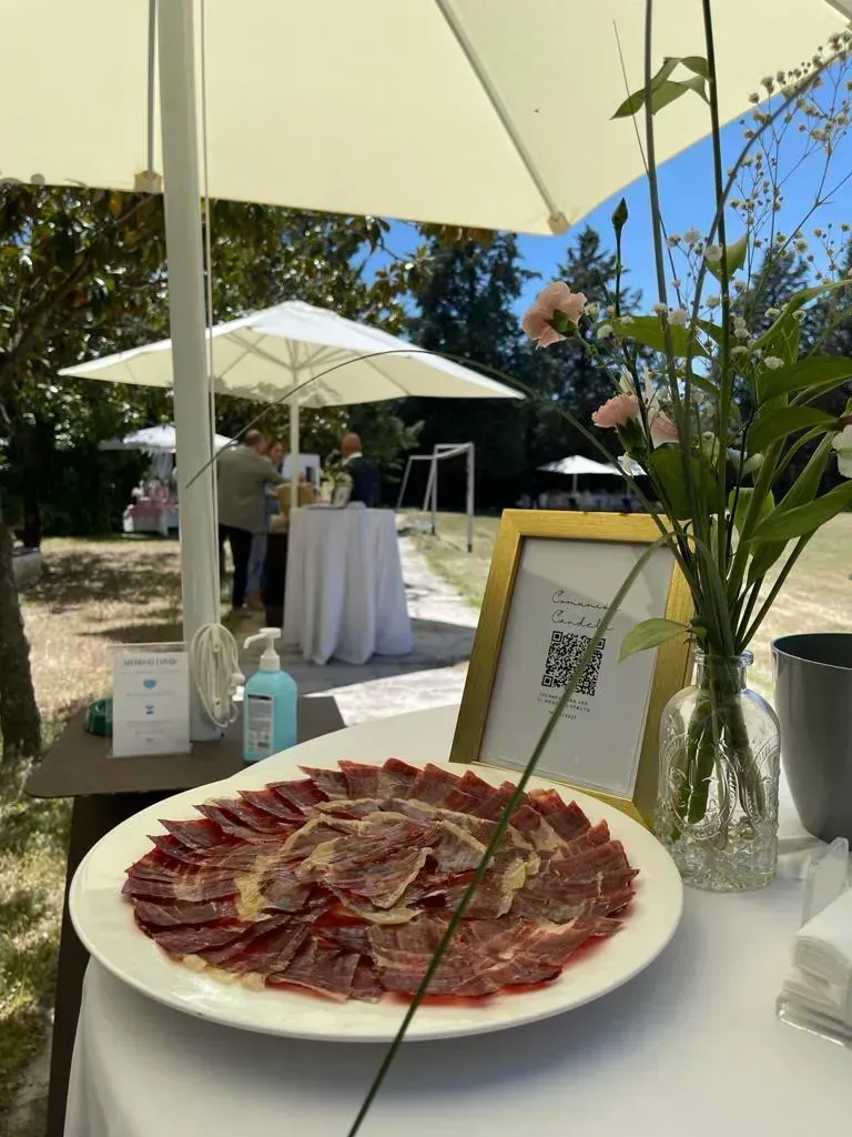 Plato de embutidos cortados en lonchas finas junto a un cartel enmarcado y un jarrón en una mesa de exterior.