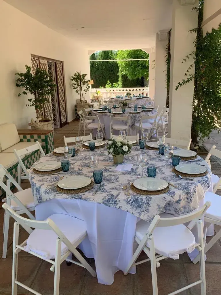 Mesa larga de banquete bajo una pérgola frondosa, con sillas doradas y vajilla blanca al aire libre.