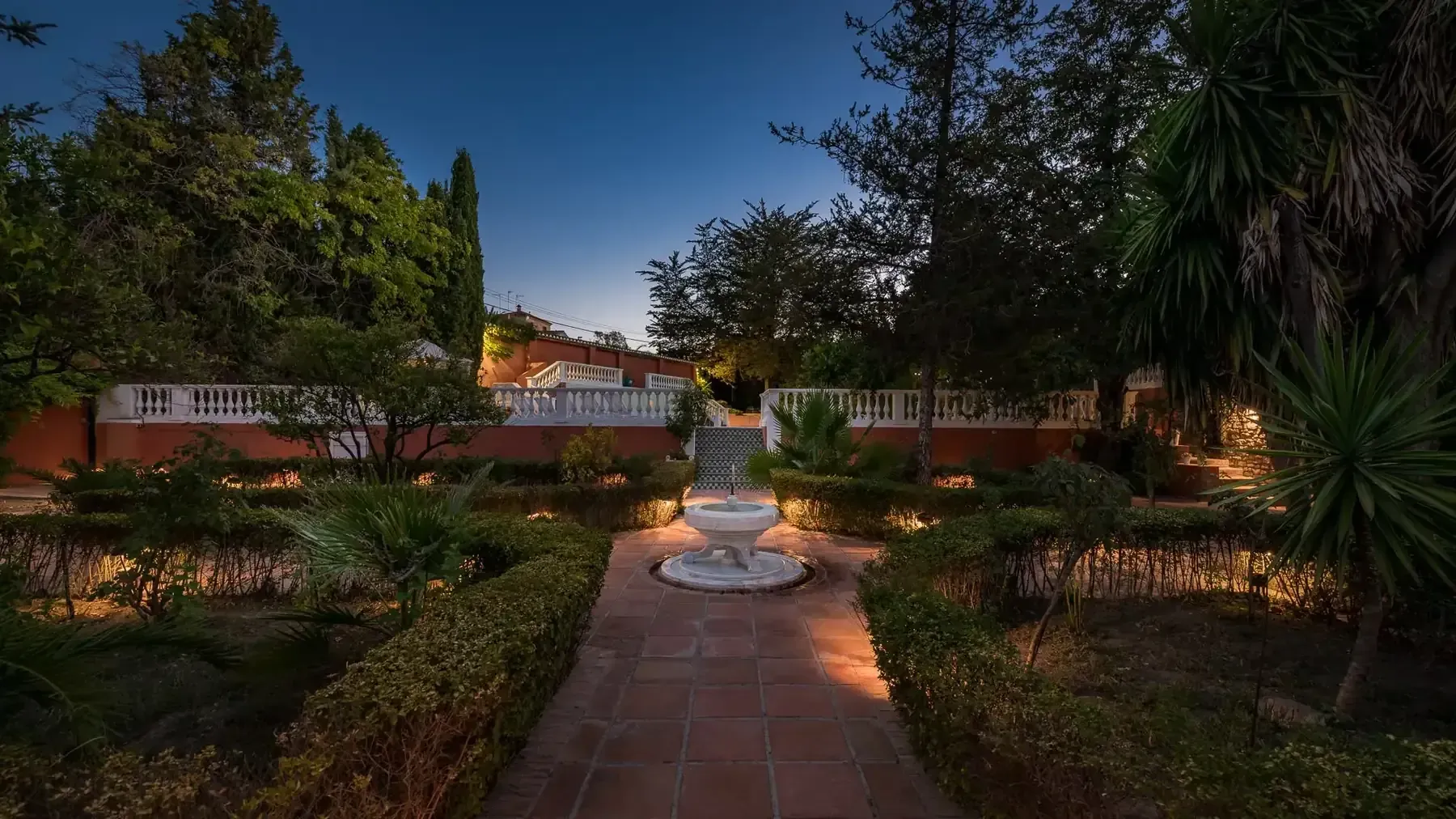 Jardín soleado con árboles, arbustos y una casa blanca al fondo.