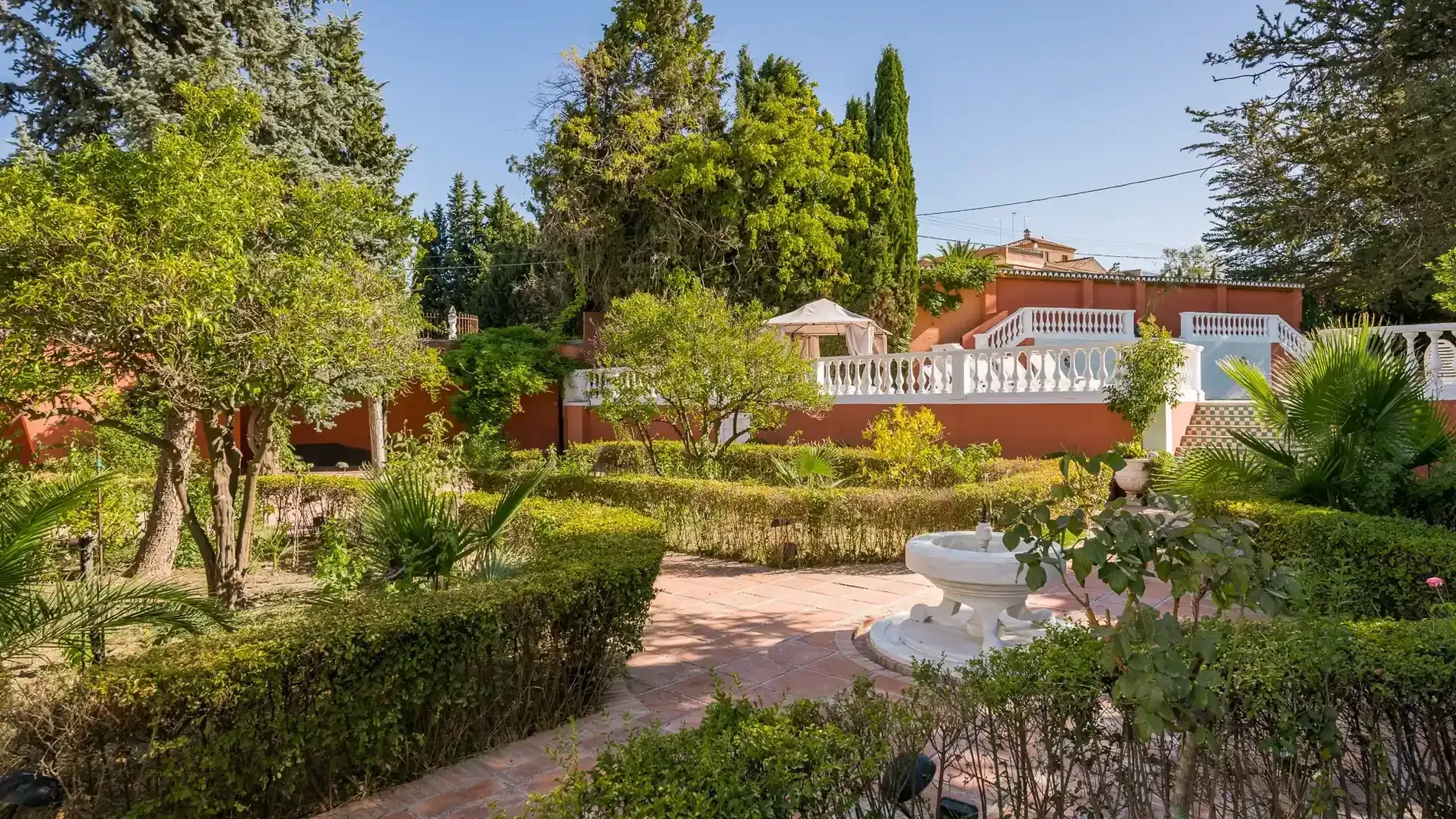 Jardín soleado con árboles, arbustos y una casa blanca al fondo.