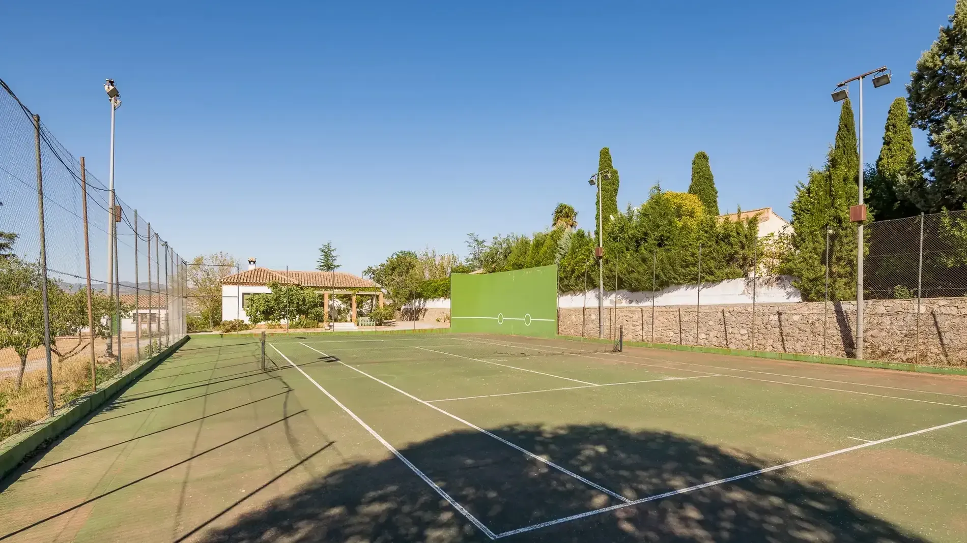 Cancha de tenis al aire libre vacía con una valla de tela metálica, paredes verdes y árboles bajo un cielo azul.