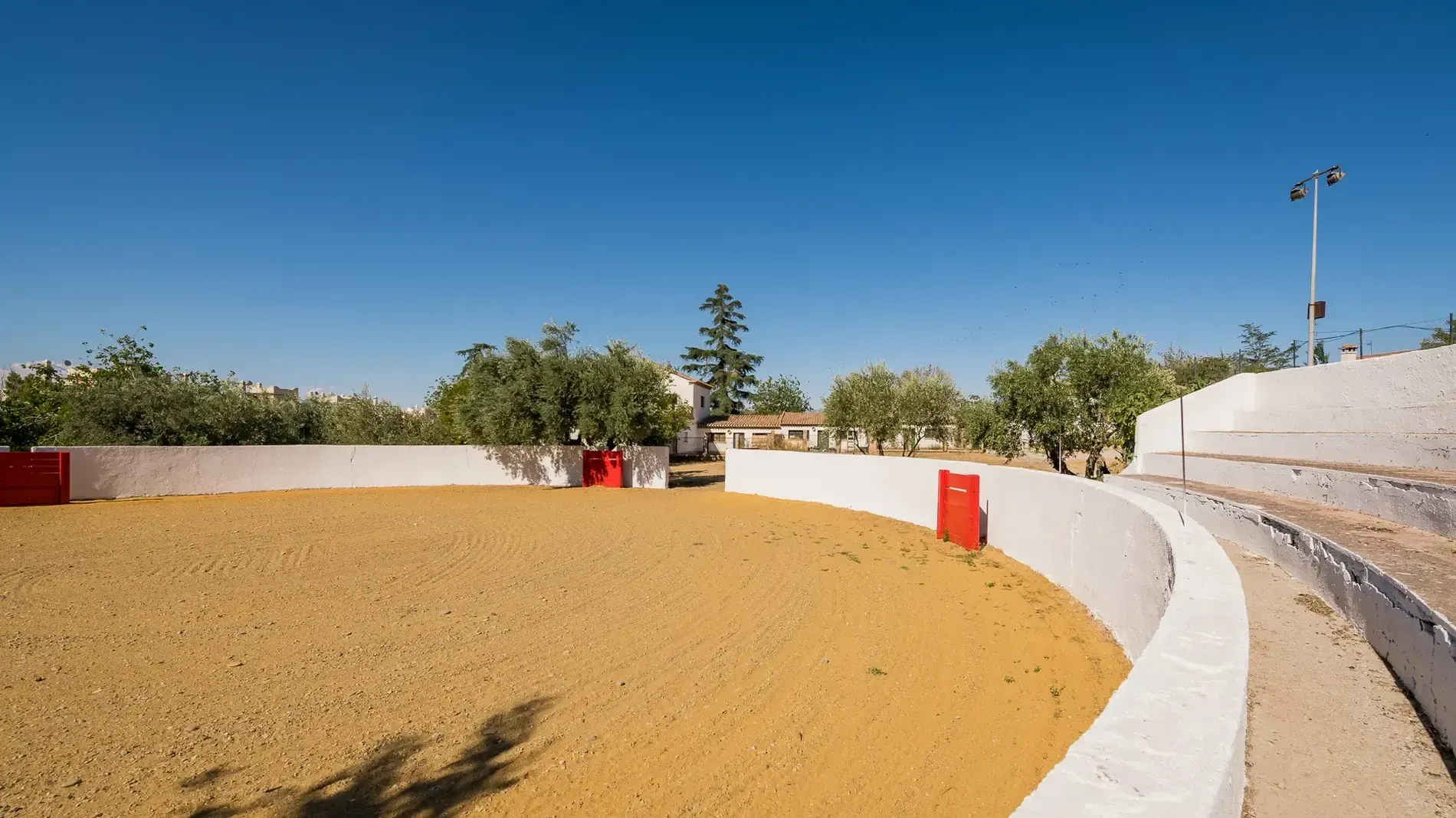 Pista de arena curva con paredes blancas bajo un cielo azul claro, con árboles y postes rojos al fondo.