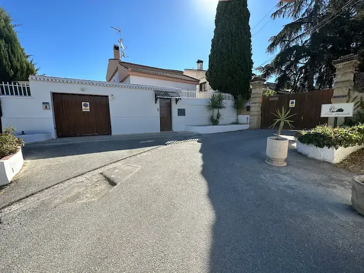 Entrada de vehículos residencial y entrada de casa blanca con garaje, árboles y cielo azul claro.