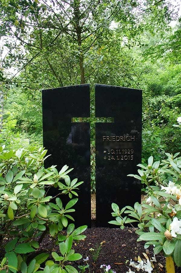 Ein schwarzer Grabstein mit einem Kreuz darauf ist von Bäumen und Blumen umgeben.