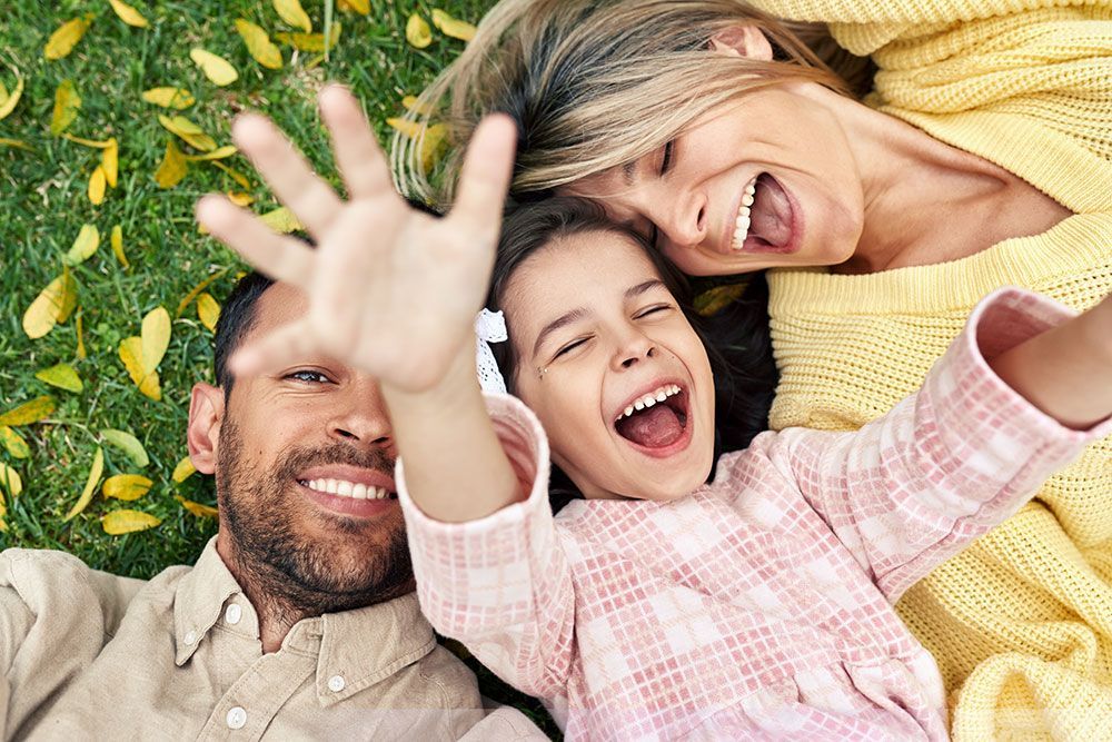 Dreiköpfige Familie liegt im Gras, lächelt und lacht und macht ein Selfie.