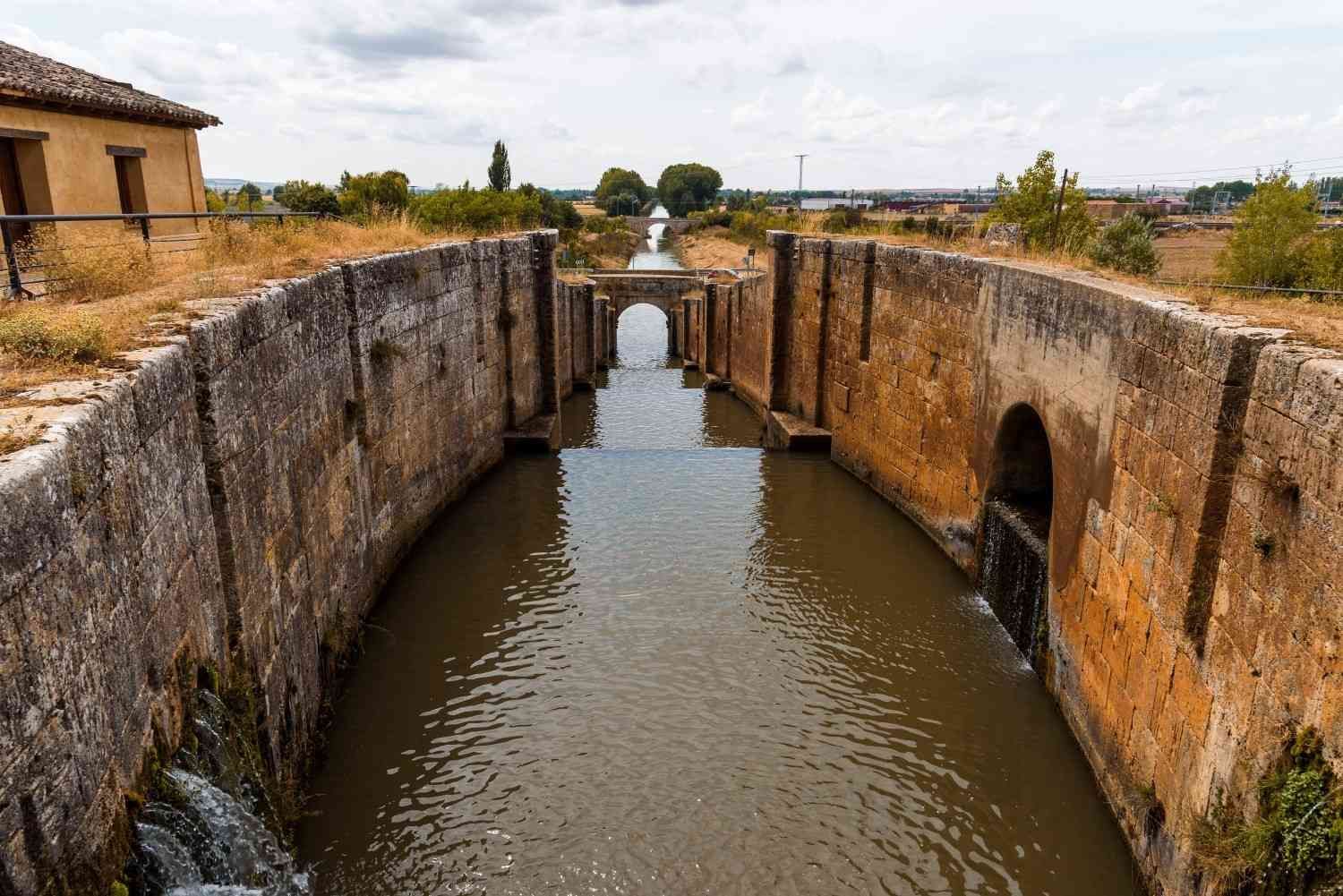Un estrecho canal de piedra entre las antiguas murallas de un fuerte, con el agua fluyendo hacia un arco lejano bajo un cielo nublado.