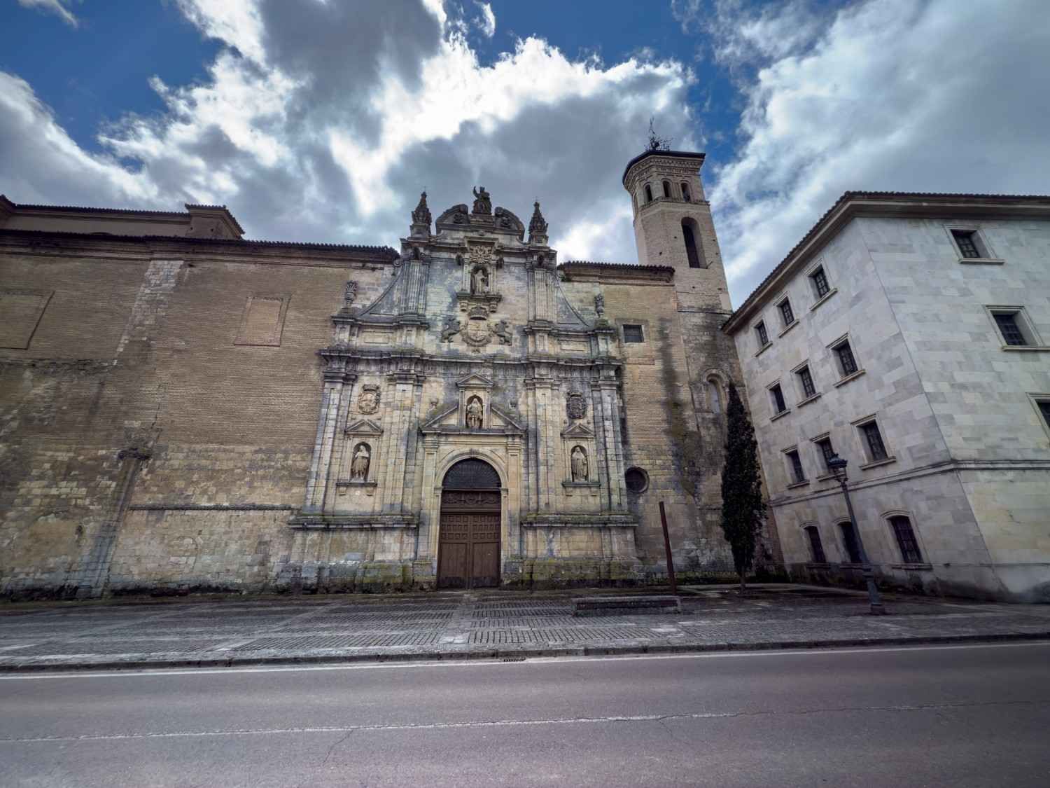 Antigua fachada de iglesia de piedra con puerta ornamentada y torre junto a un gran muro desgastado bajo un cielo nublado.