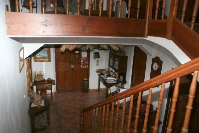Vista interior del vestíbulo de una casa con escalera de madera, barandilla de balcón y suelo de baldosas.
