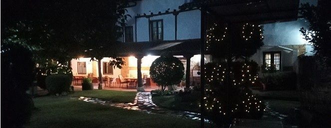 Exterior de la casa iluminado por la noche con luces en el porche, guirnaldas de luces y arbustos decorados en el jardín.