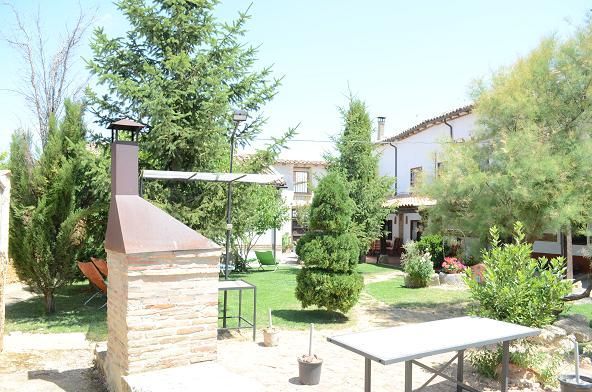 Patio trasero soleado con parrilla de ladrillo, mesa, sillas y frondosos árboles y arbustos verdes.