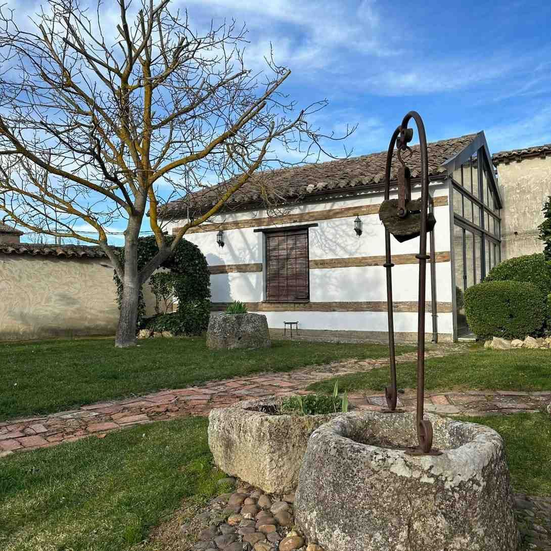 Patio rústico con pozo de piedra, árbol y edificio encalado bajo un cielo azul.