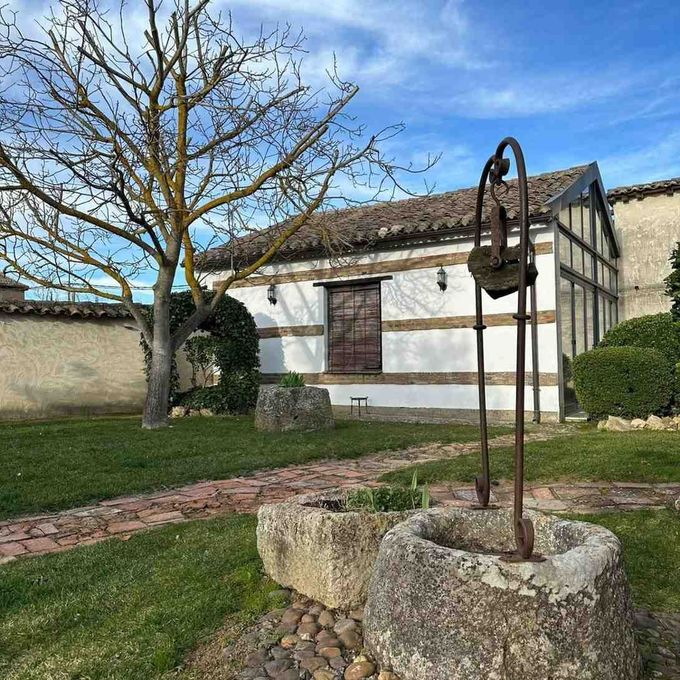 Patio rústico con pozo de piedra, árbol y edificio encalado bajo un cielo azul.