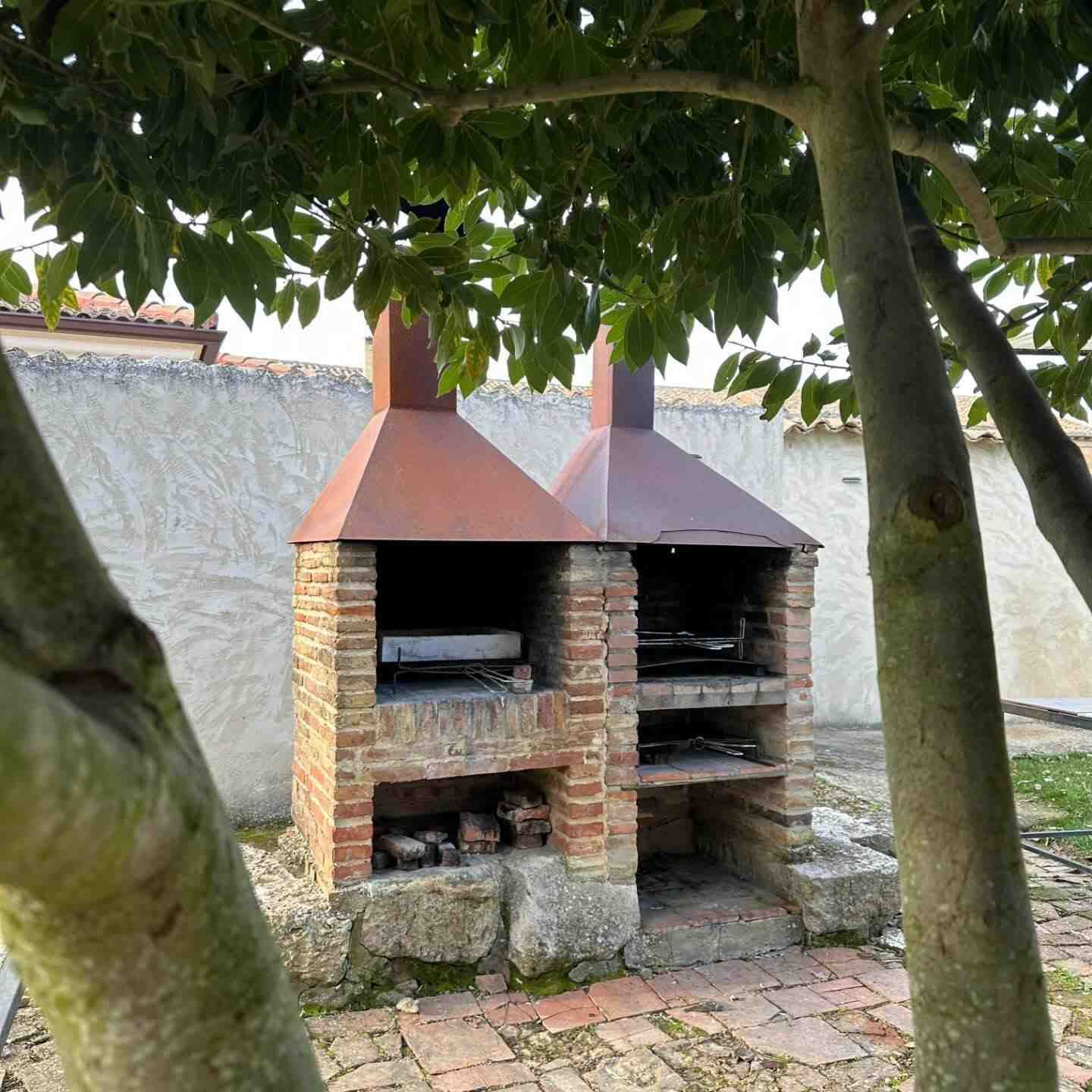 Parrilla de ladrillo al aire libre con dos chimeneas de color óxido bajo un árbol, junto a un muro de piedra y un patio pavimentado.