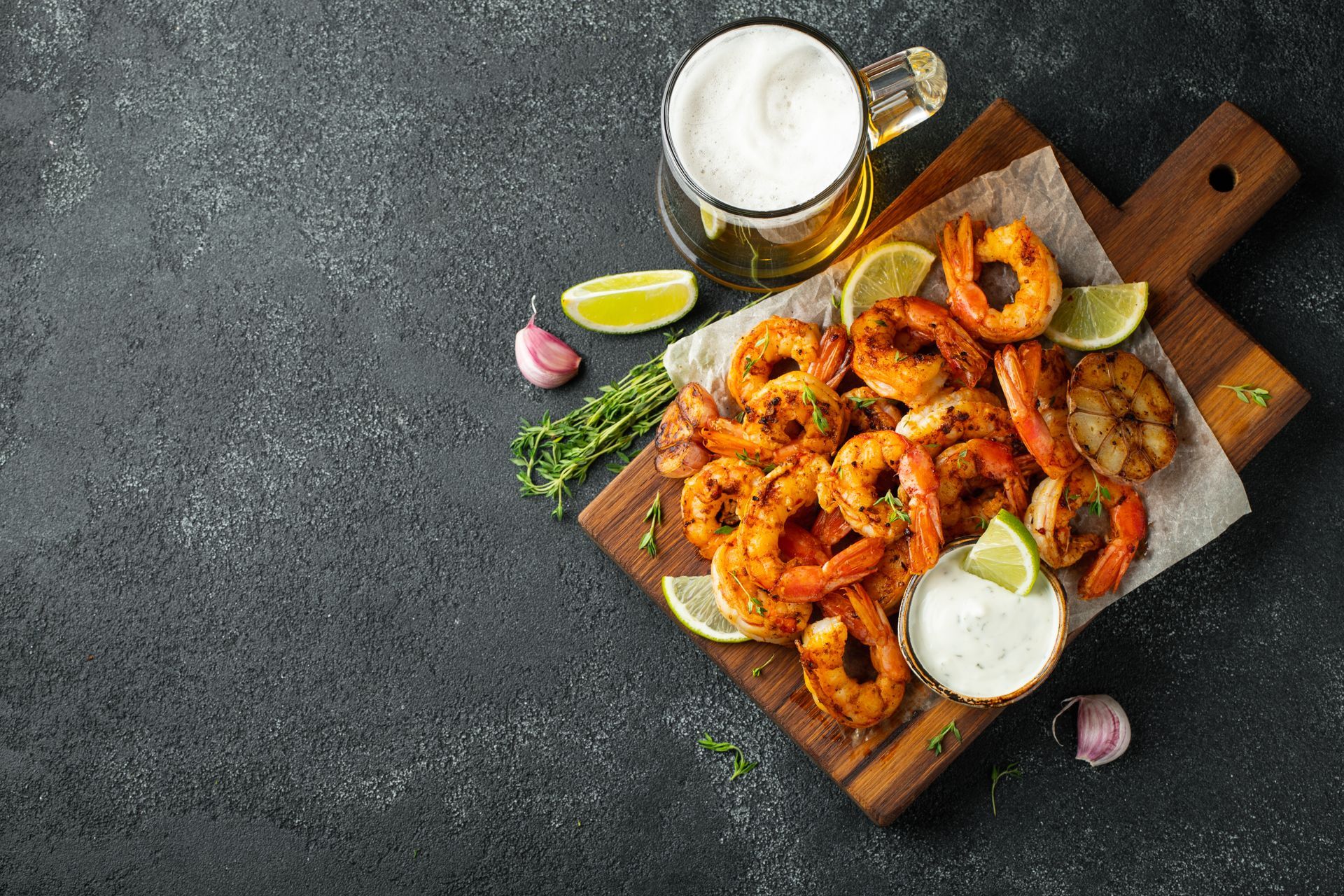 Crevettes et sauce à tremper sur une planche en bois, avec du citron vert et des herbes, sur une table sombre