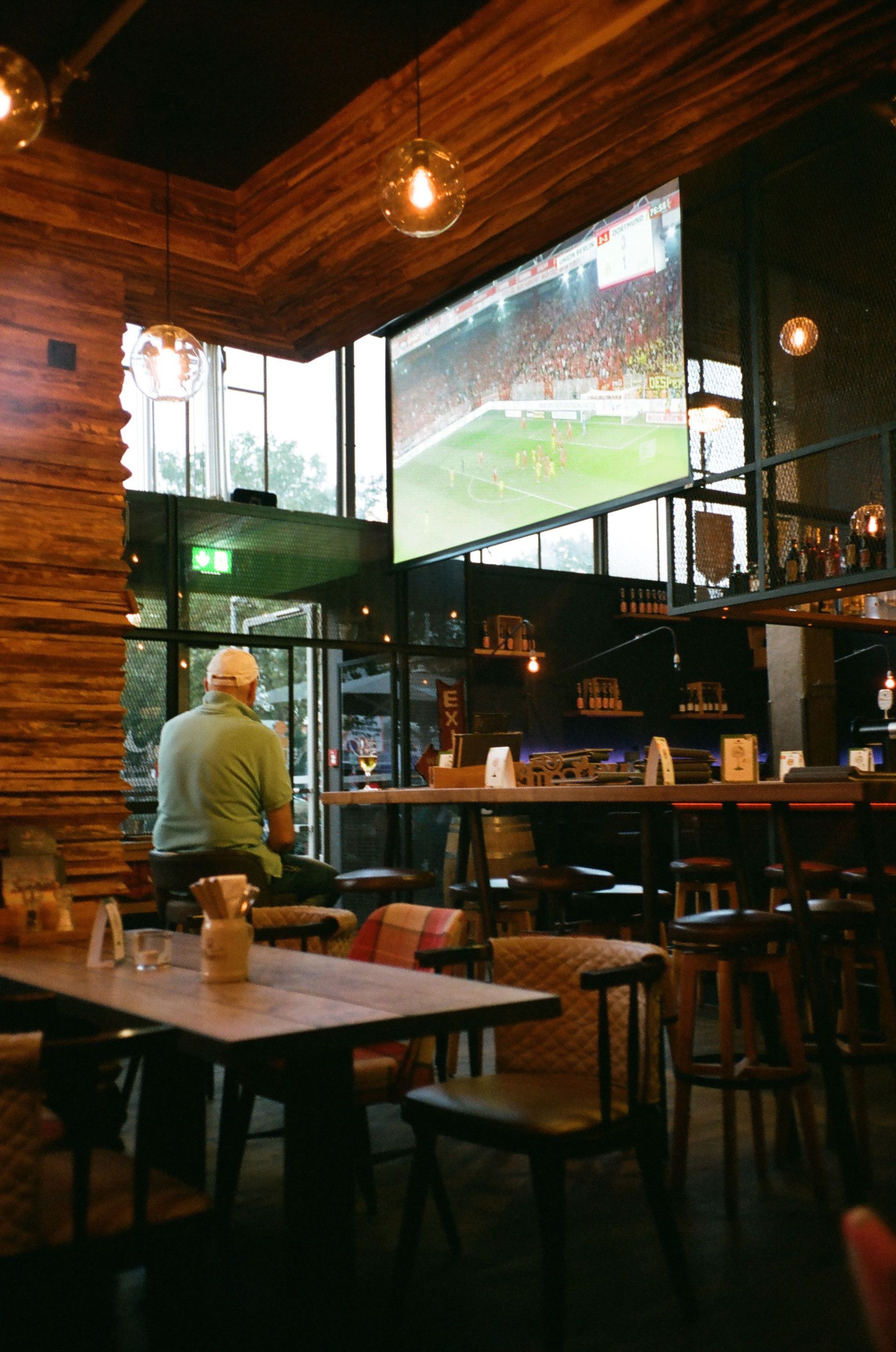 Intérieur chaleureux d’un bar avec un grand écran de télévision diffusant un match de football et une personne qui le regarde près du comptoir