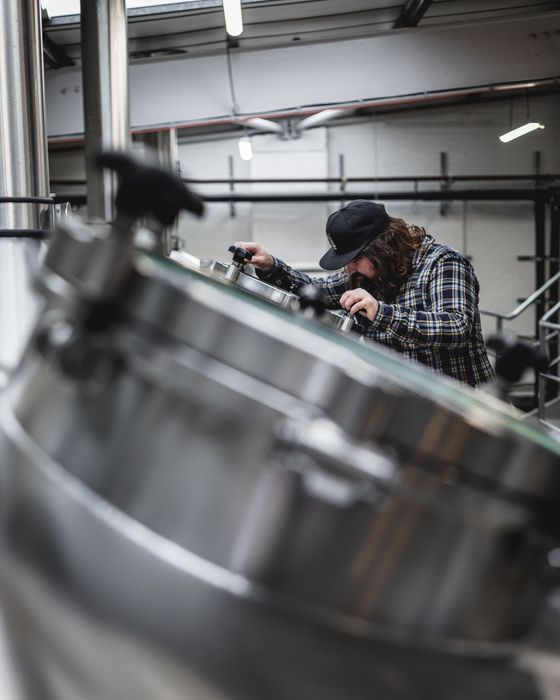 Un homme en chemise à carreaux et casquette dans une brasserie, regarde dans une cuve métallique.