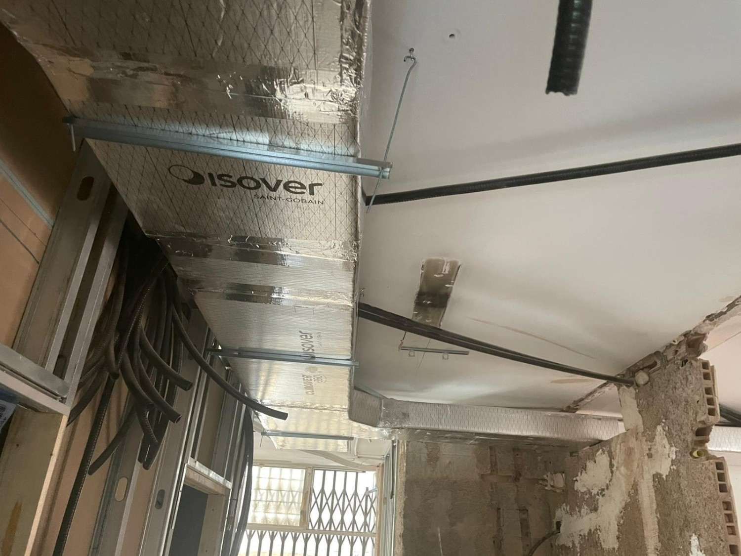 Techo dañado con conductos de ventilación plateados expuestos y aislamiento colgante en un pasillo interior sin terminar.