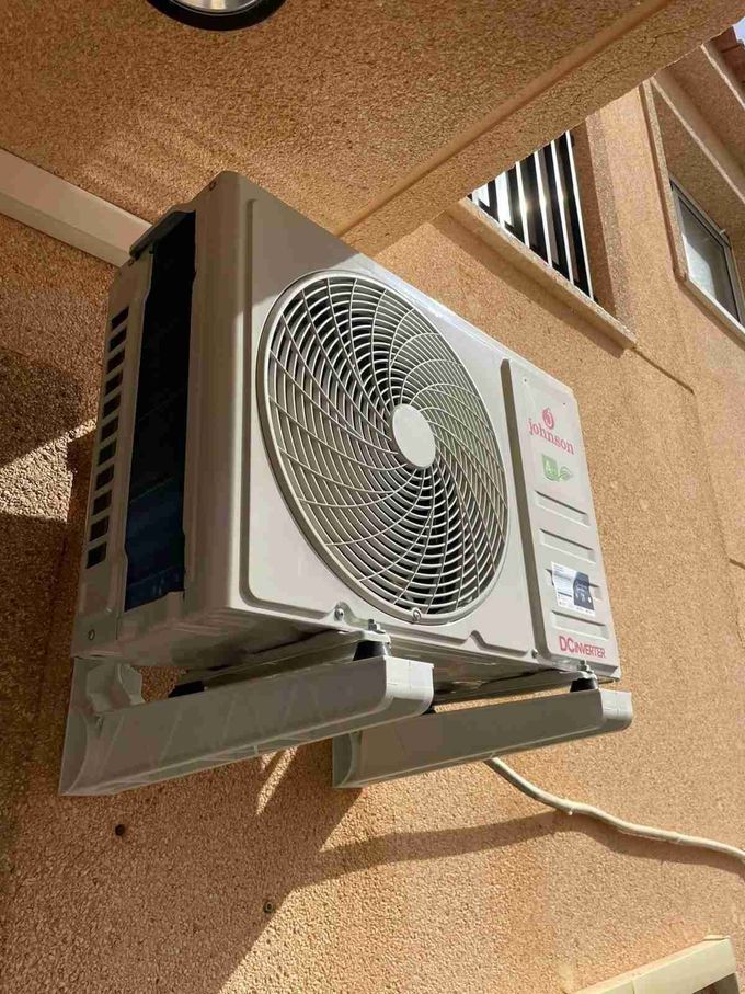 Unidad de aire acondicionado exterior montada en la pared, sobre la fachada de estuco color beige de un edificio.