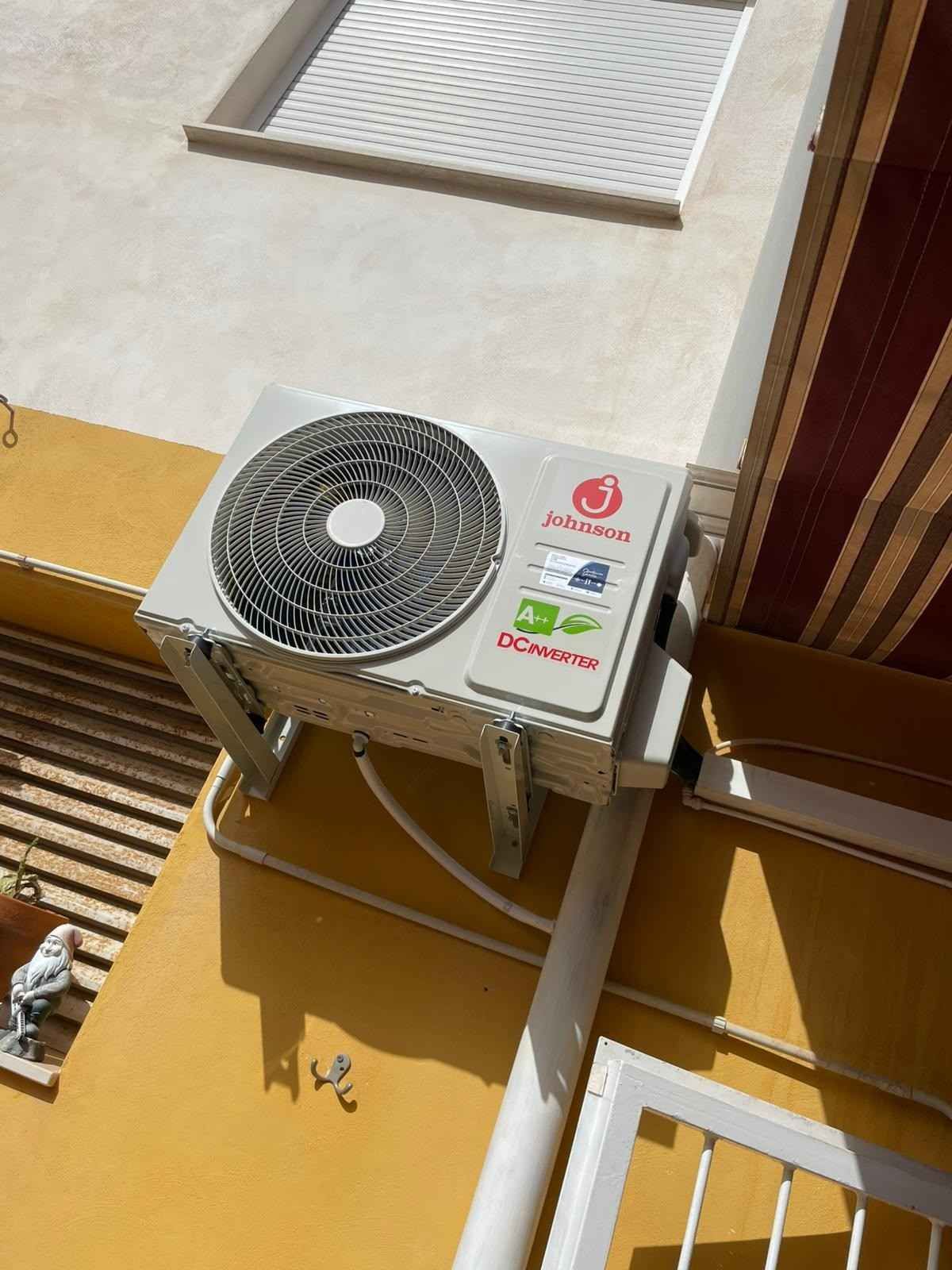 Unidad exterior de aire acondicionado montada en una pared amarilla debajo de una ventana, con la rejilla del ventilador visible.