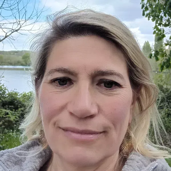 Les mains jointes sur les genoux, elle porte une chemise crème à manches longues. Assise devant un bloc-notes, on aperçoit sa jupe verte.