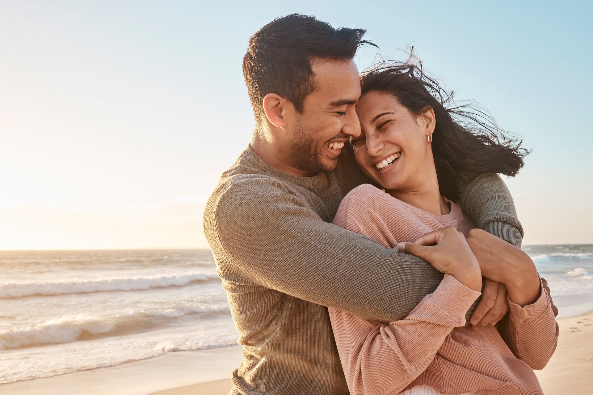 Un couple s'enlace et sourit sur une plage ensoleillée, avec les vagues et le ciel en arrière-plan.