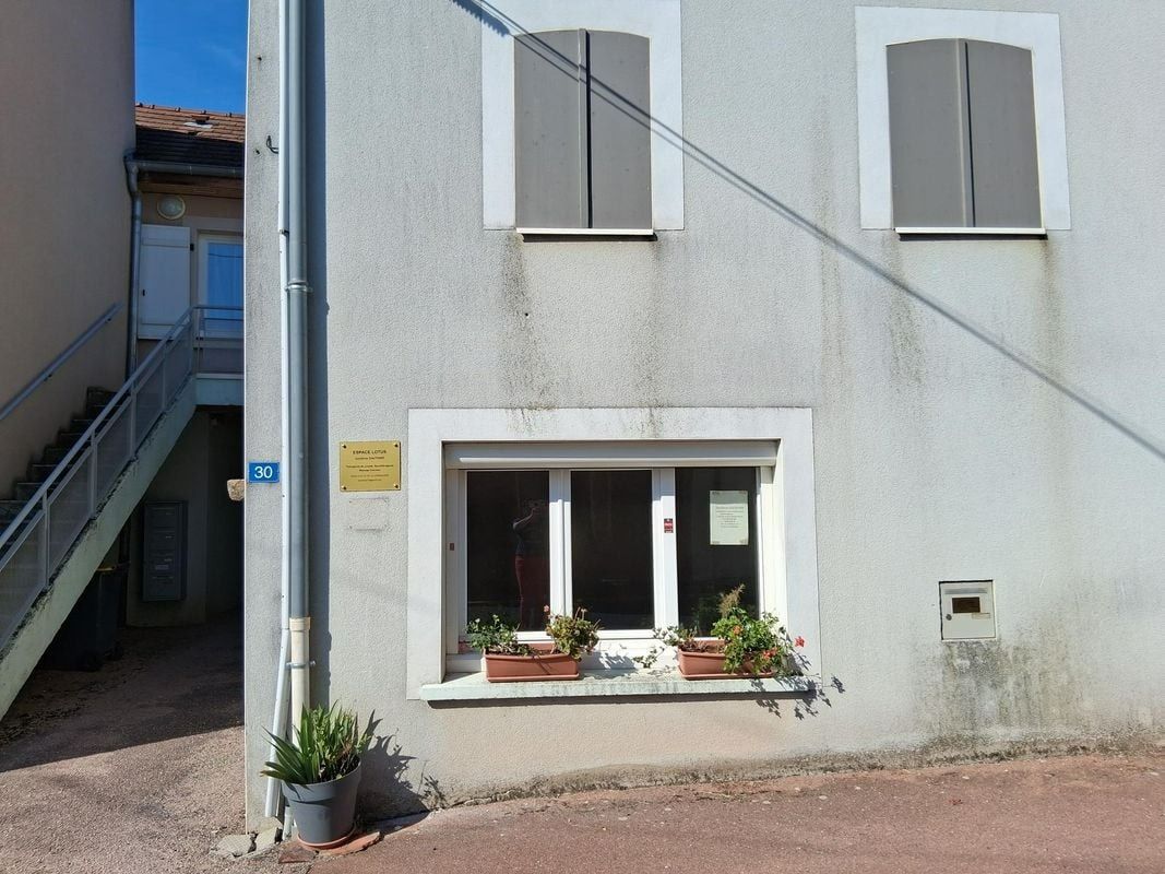 Bâtiment gris avec trois fenêtres, volets gris et une fenêtre plus basse avec des plantes en pot. Escalier sur le côté.
