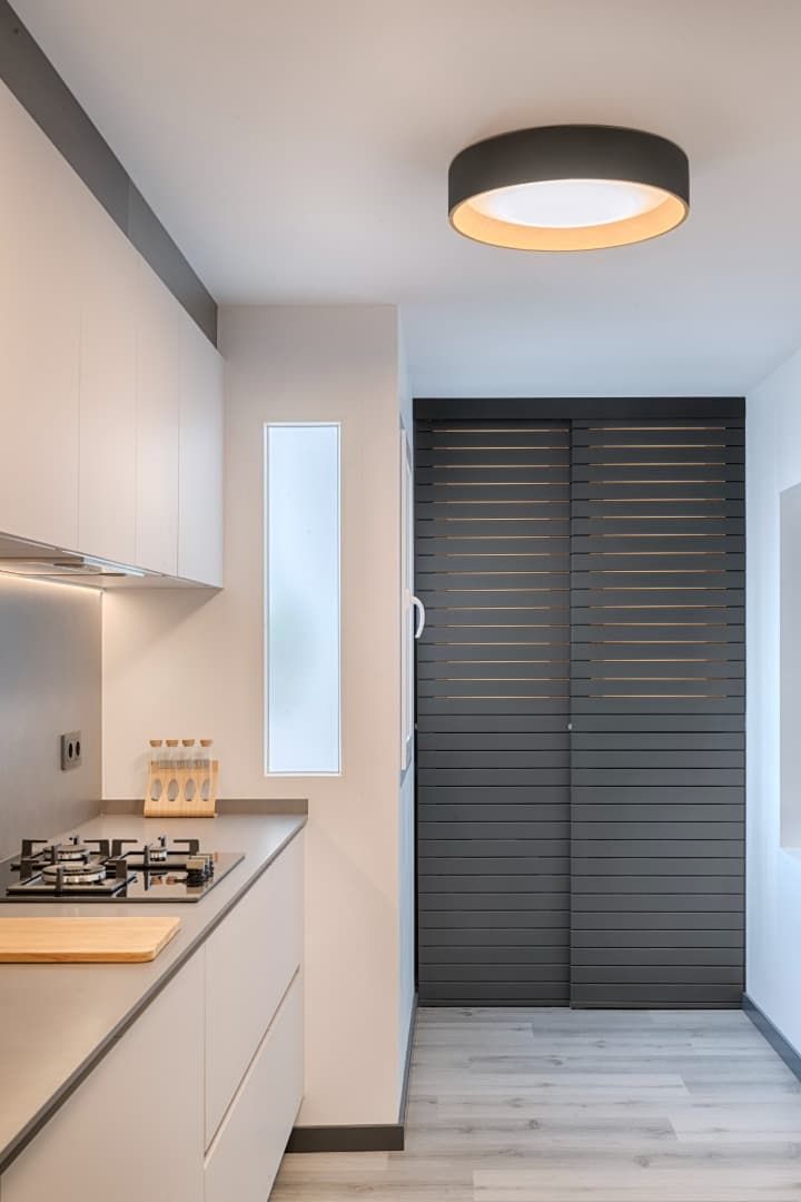 Cocina moderna y estrecha con armarios blancos, suelo de madera clara y puertas correderas oscuras en los armarios.