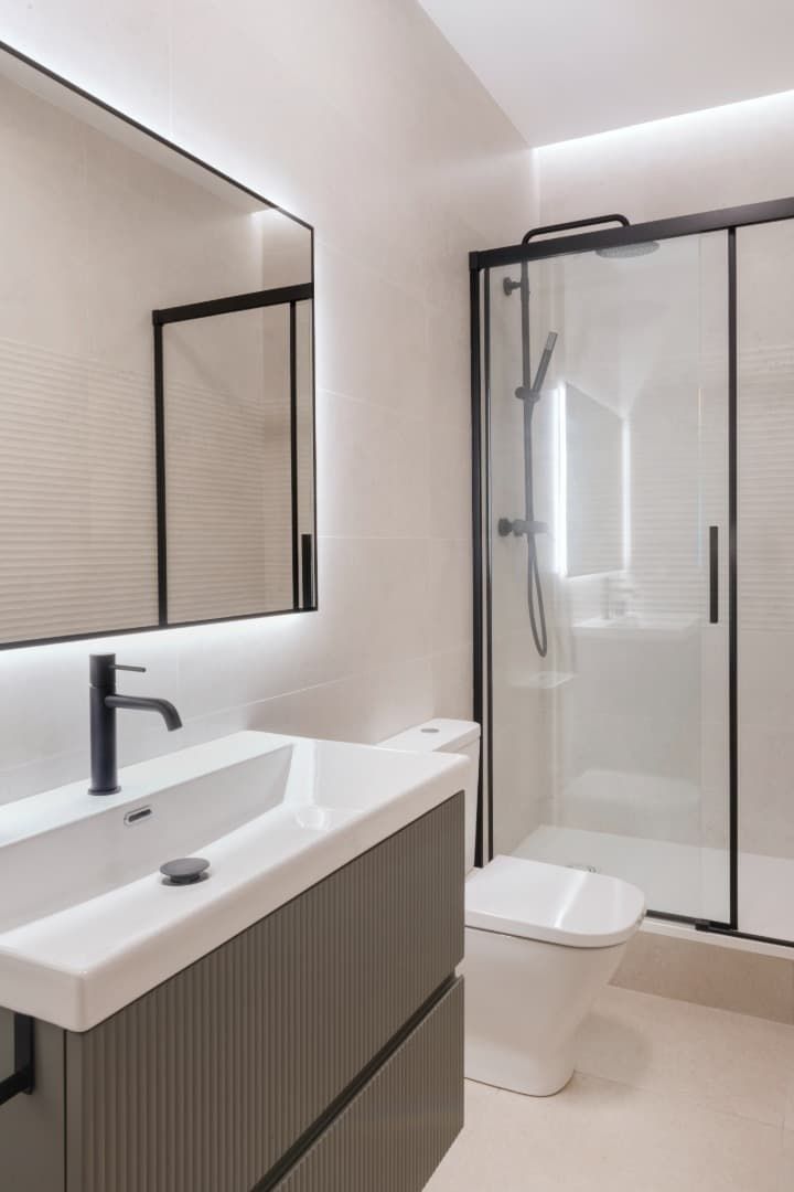 Baño moderno con espejo de pared, lavabo blanco, inodoro y cabina de ducha de cristal.