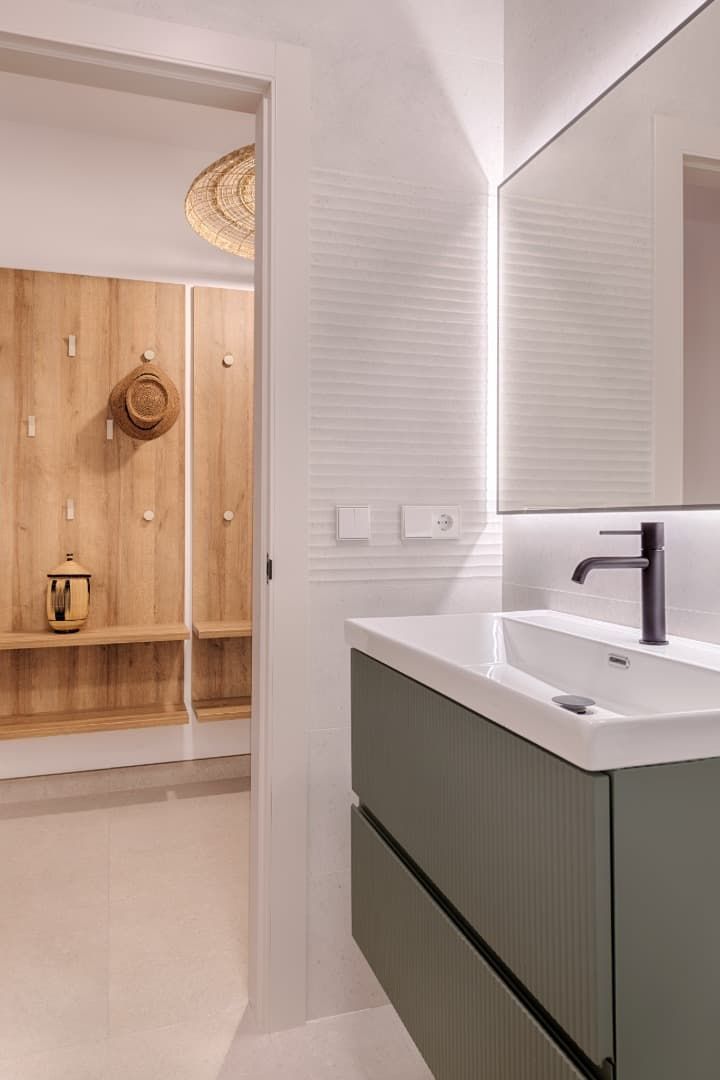 Tocador de baño moderno con lavabo blanco, grifo negro y espejo grande; al fondo se ve un pasillo con un banco de madera.