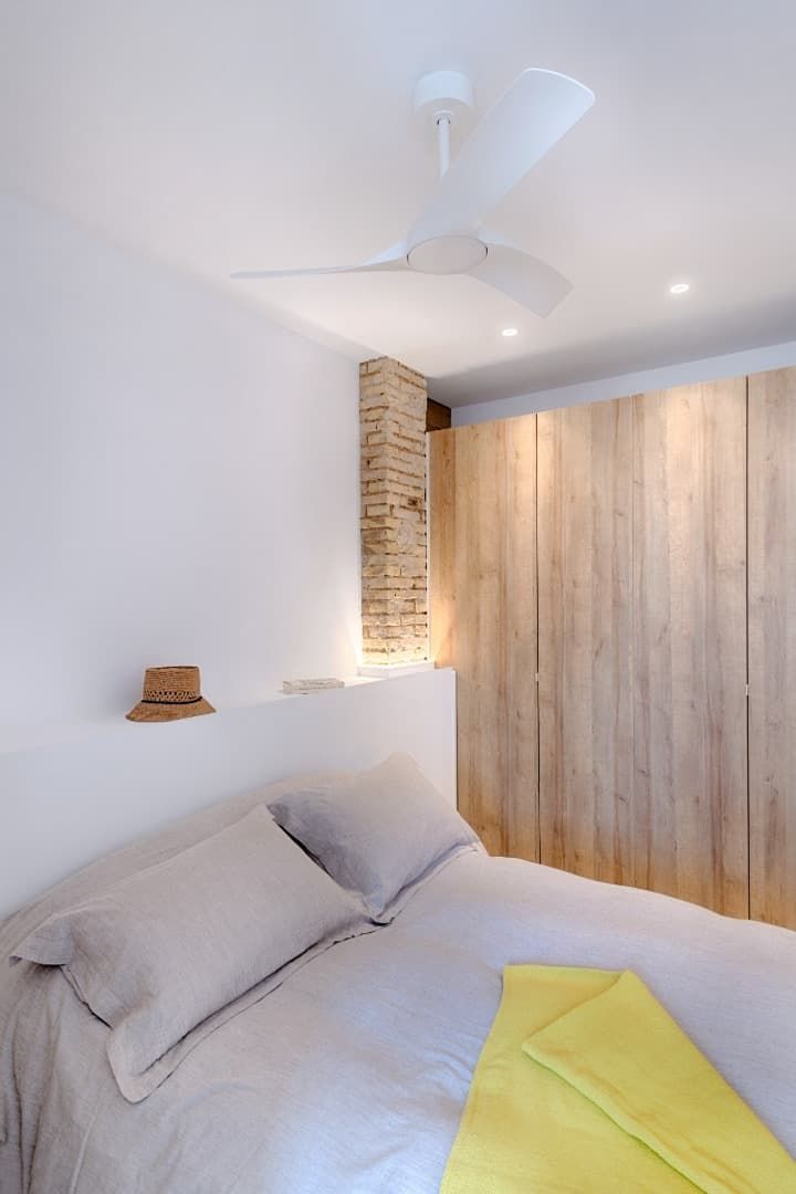 Dormitorio luminoso y minimalista con cama blanca, pared decorativa de madera y ventilador de techo.