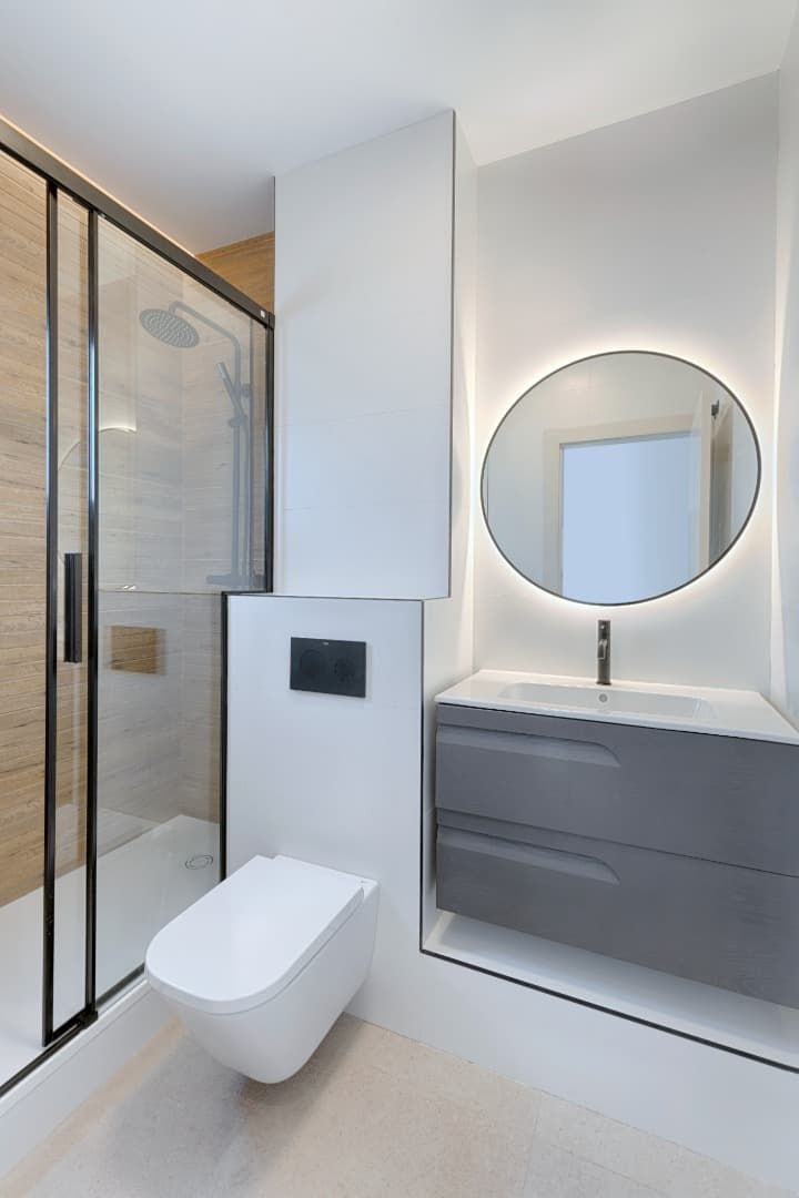 Baño moderno con ducha de cristal, inodoro suspendido y espejo redondo retroiluminado sobre un tocador gris.