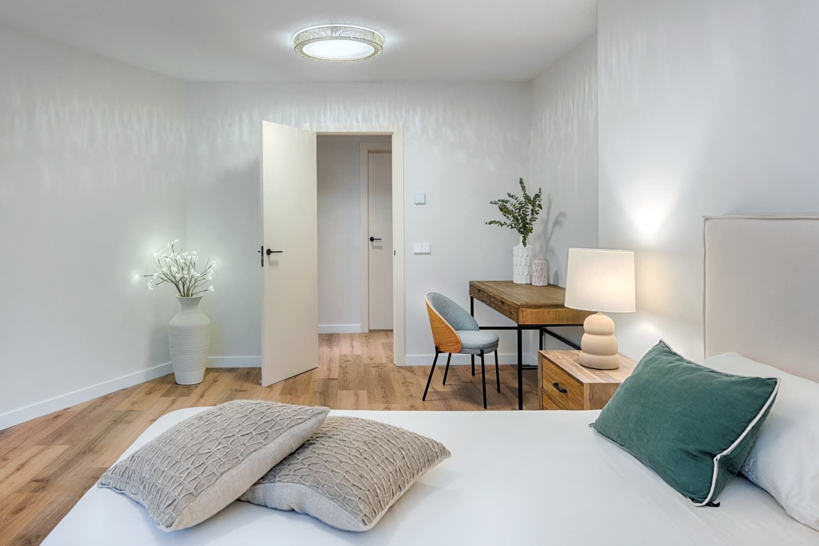 Habitación luminosa con paredes blancas, suelo de madera, cama, escritorio, lámpara y plantas en macetas.