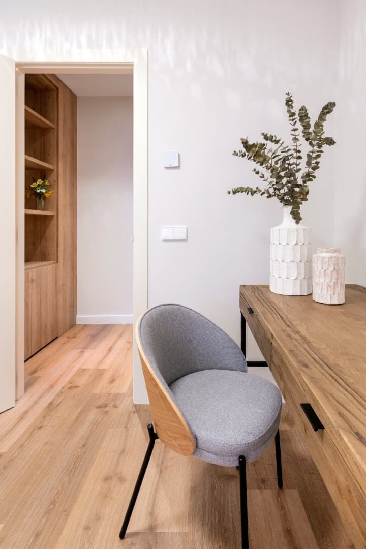 Comedor minimalista con mesa de madera, silla gris y jarrón blanco con ramas junto a una puerta abierta.