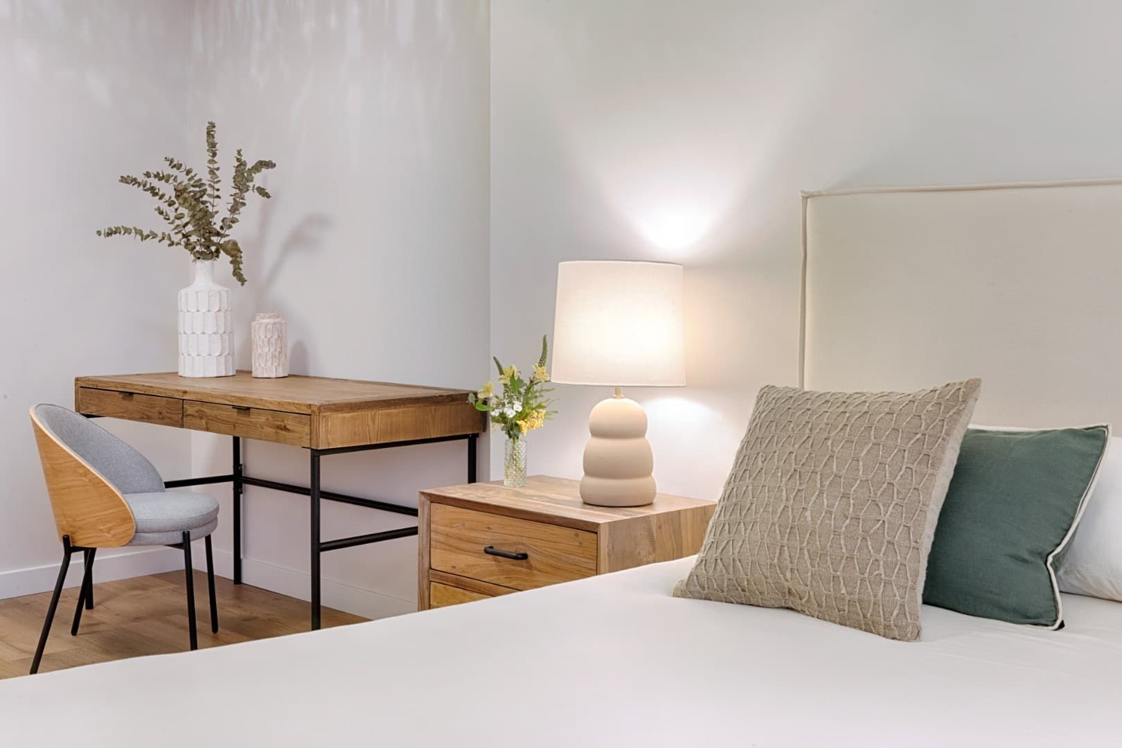 Dormitorio luminoso y minimalista con cama blanca, escritorio de madera, lámpara y decoración neutra.