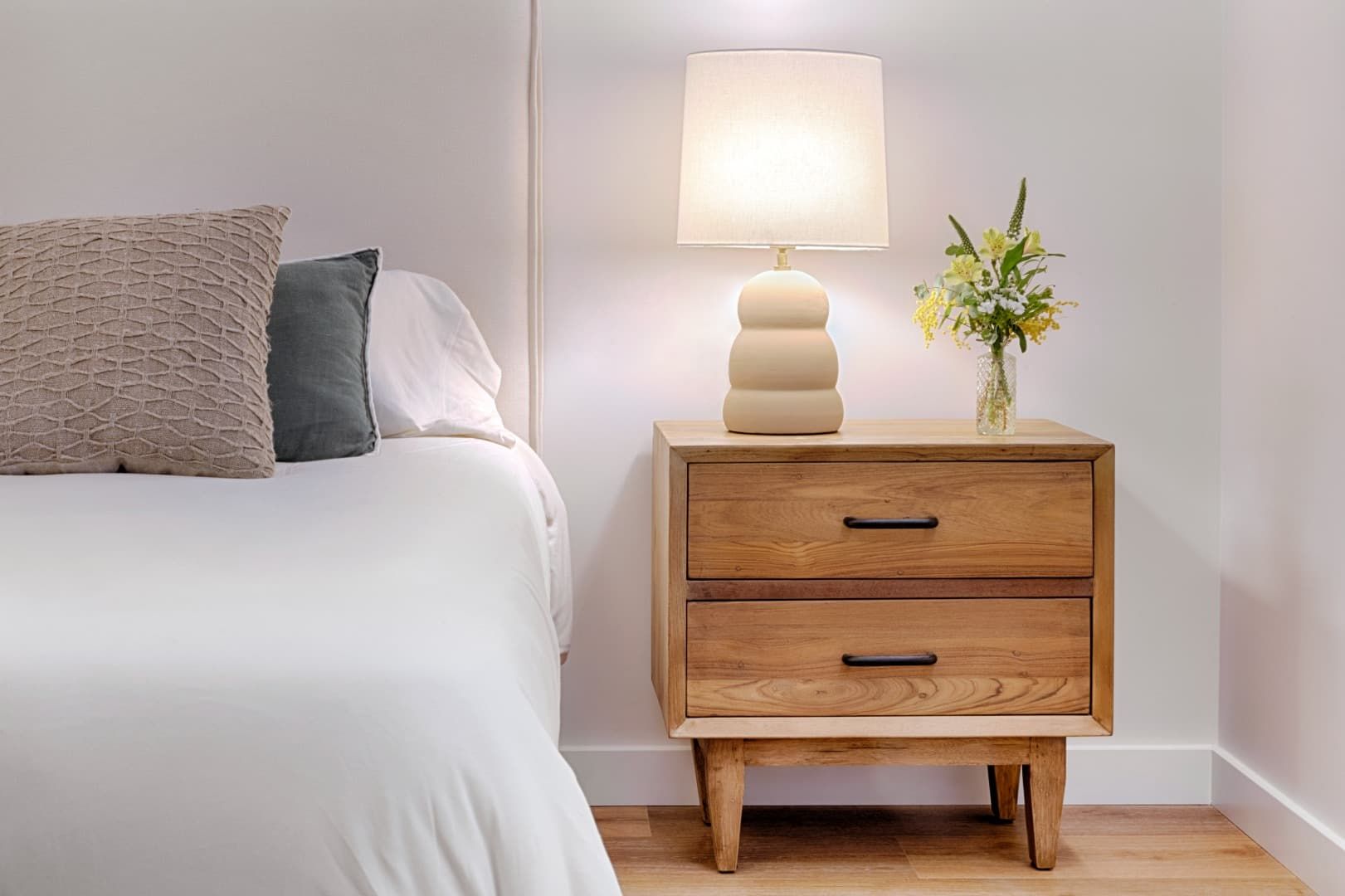 Dormitorio minimalista con cama blanca, mesita de noche de madera, lámpara y un pequeño jarrón con flores.