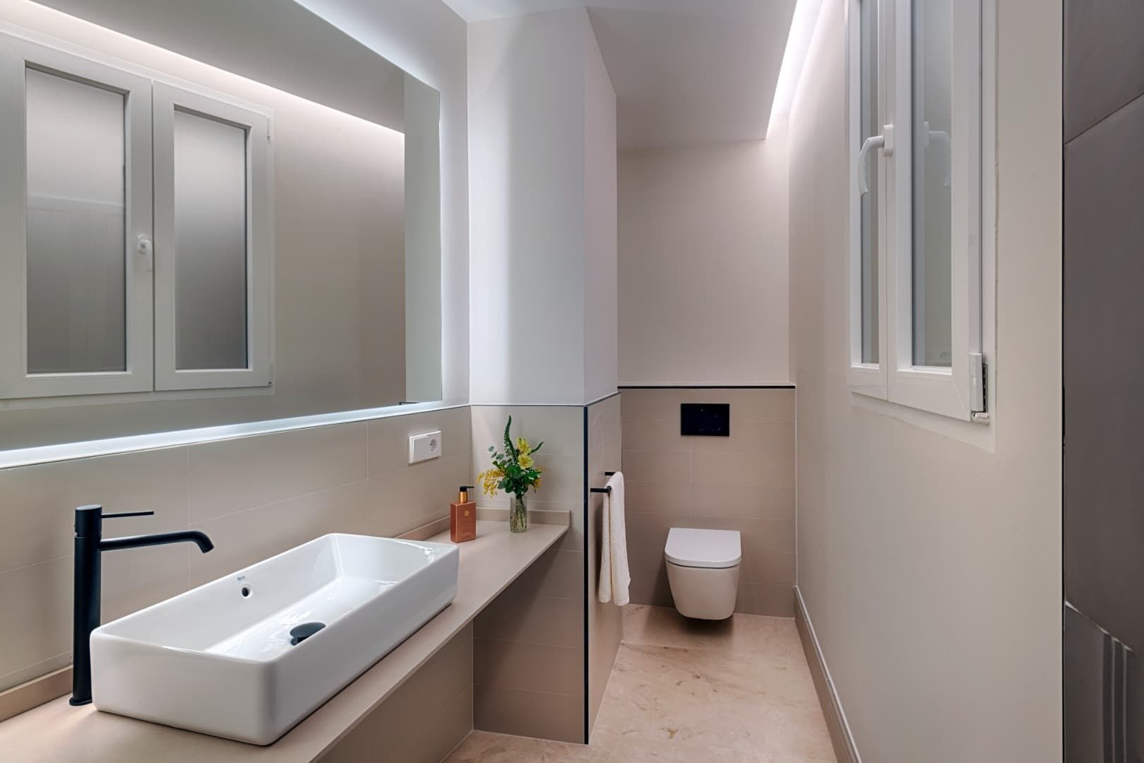 Baño moderno de color beige con lavabo mural, espejo, inodoro y ventana estrecha.