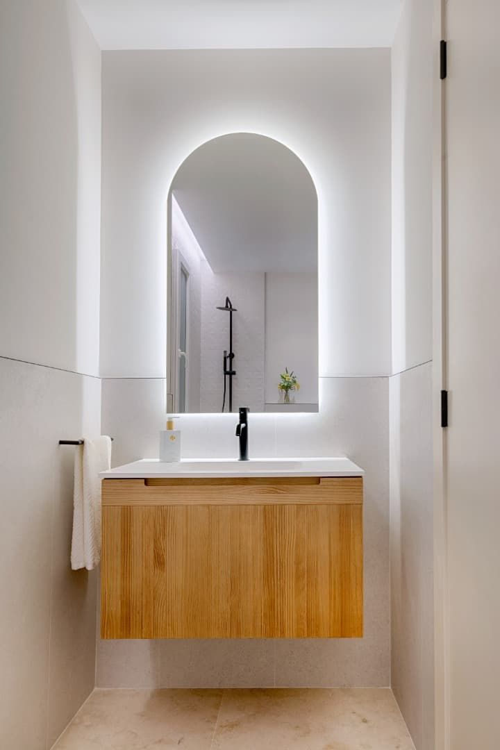 Tocador de baño moderno con mueble de madera, lavabo blanco y espejo arqueado en un interior luminoso y minimalista.