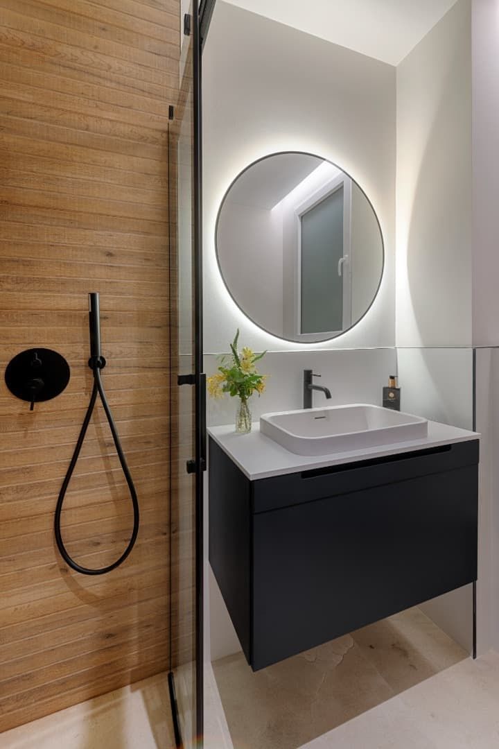 Tocador de baño moderno con espejo redondo, mueble de lavabo negro y pared de ducha revestida de madera con cabezal de ducha de mano.