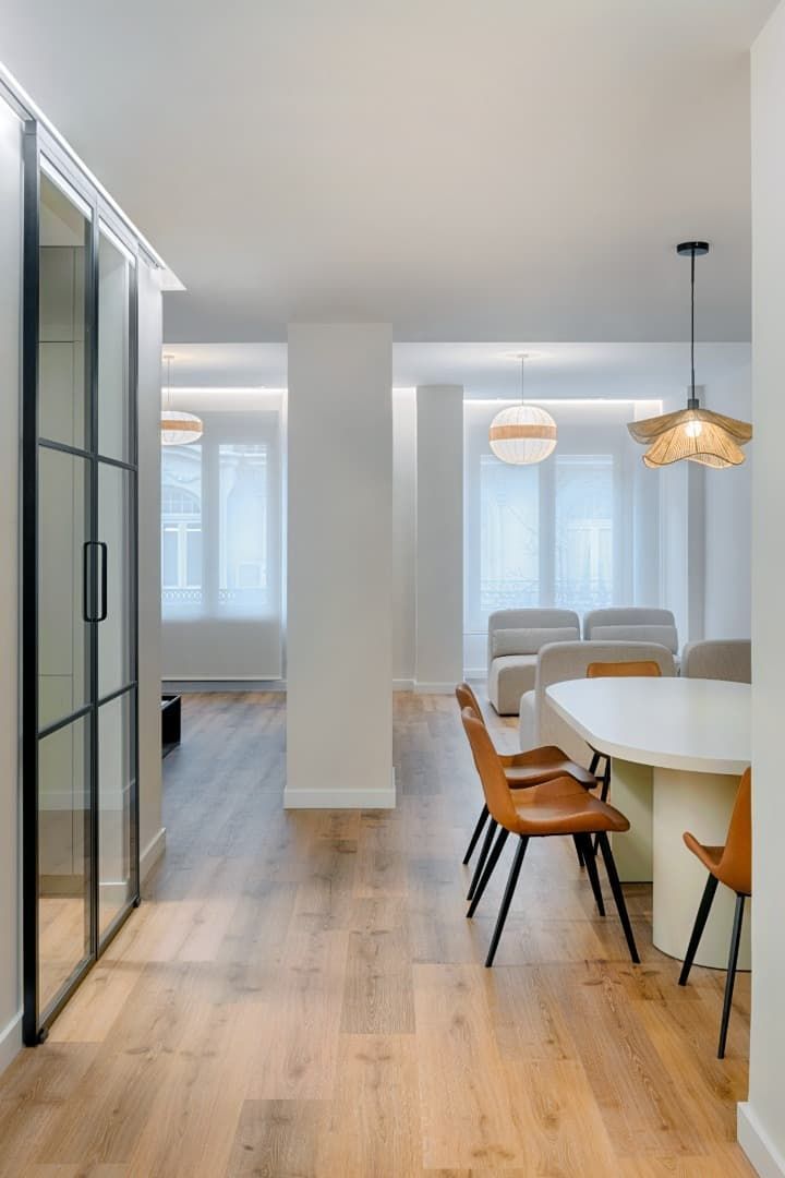 Comedor luminoso y moderno con paredes blancas, suelos de madera, mesa redonda y lámparas colgantes tejidas.