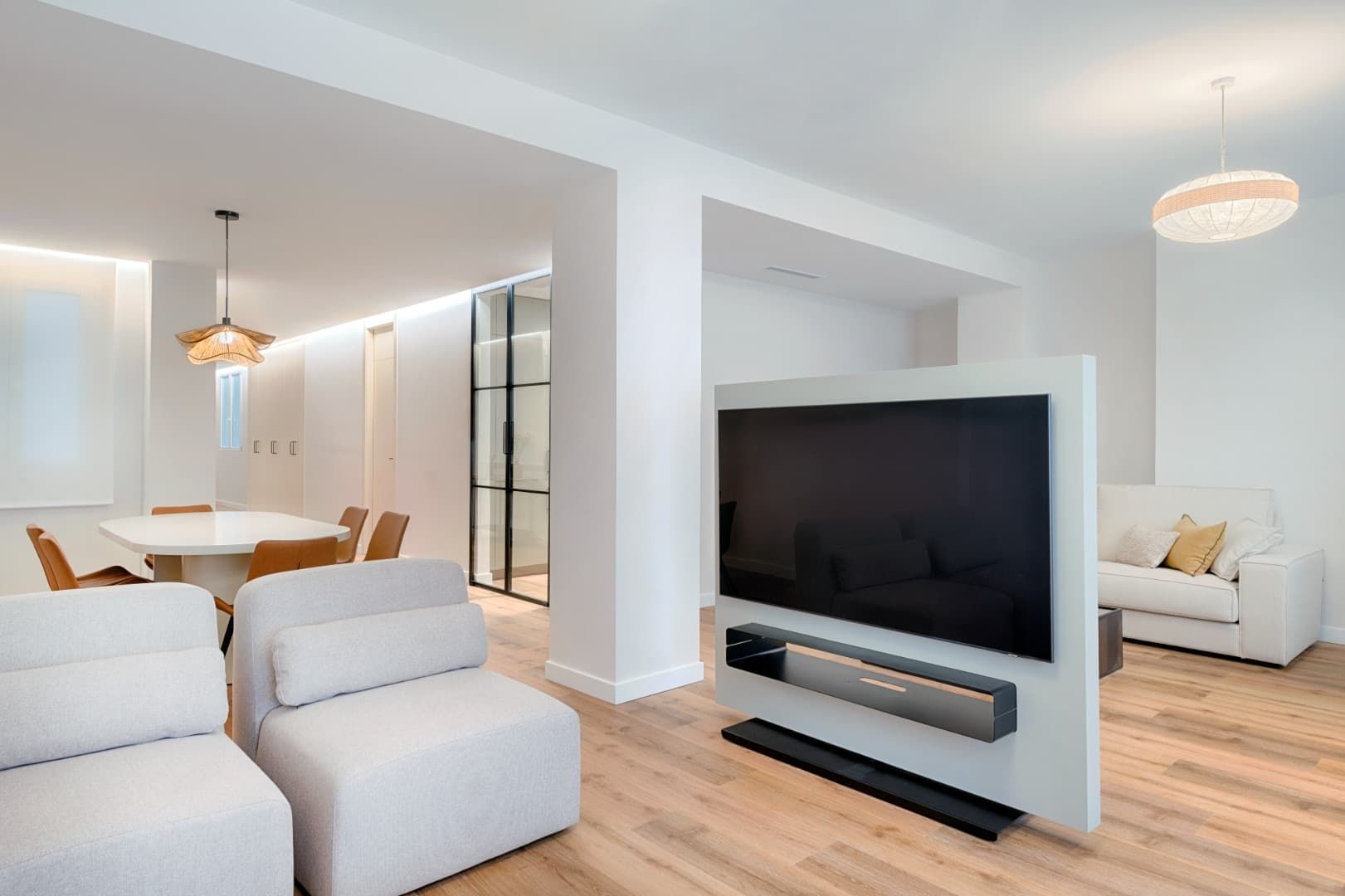 Salón-comedor luminoso y diáfano con muebles blancos, suelos de madera y un televisor grande en una mampara.