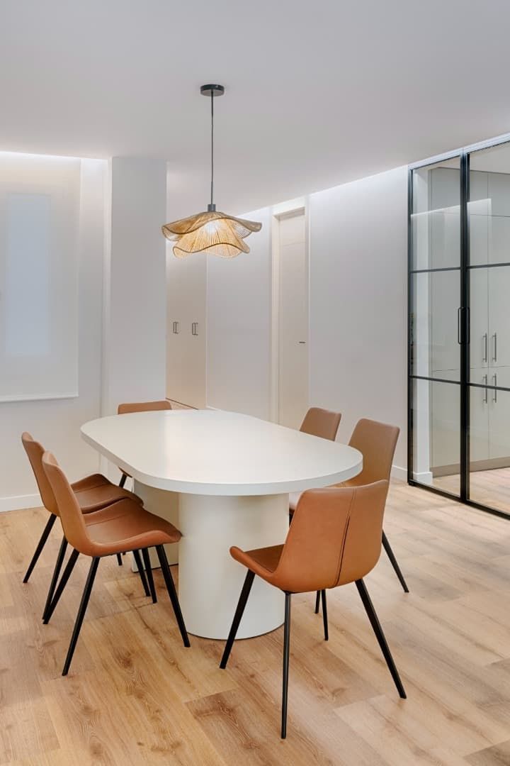Comedor moderno con mesa ovalada blanca, sillas color canela, lámpara colgante y mampara de cristal.