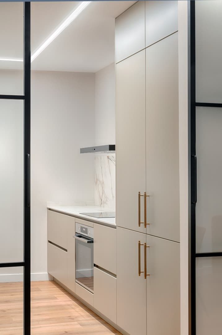 Cocina moderna de color beige con armarios empotrados, horno de acero inoxidable y suelo de madera clara, vista a través de una puerta.
