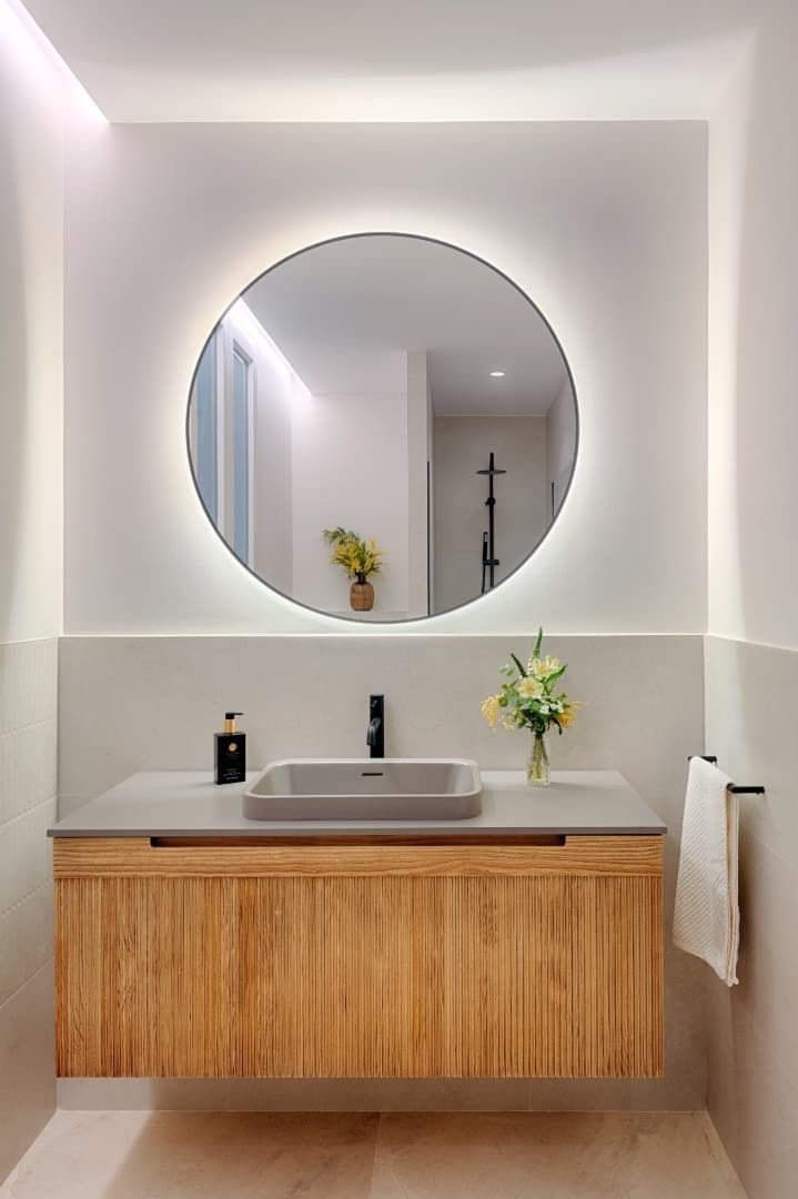 Tocador de baño moderno con espejo redondo, mueble de madera y lavabo blanco; toalla y flores sobre la encimera.