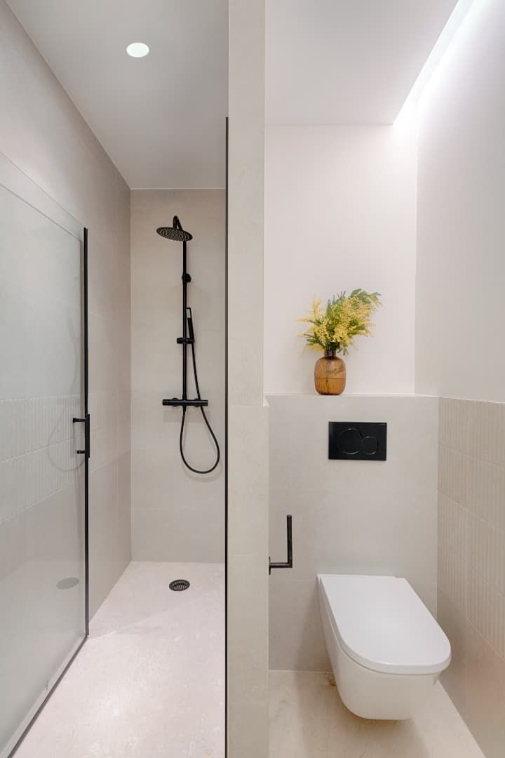Baño moderno con ducha a ras de suelo, inodoro suspendido y un pequeño jarrón de flores sobre una repisa estrecha.