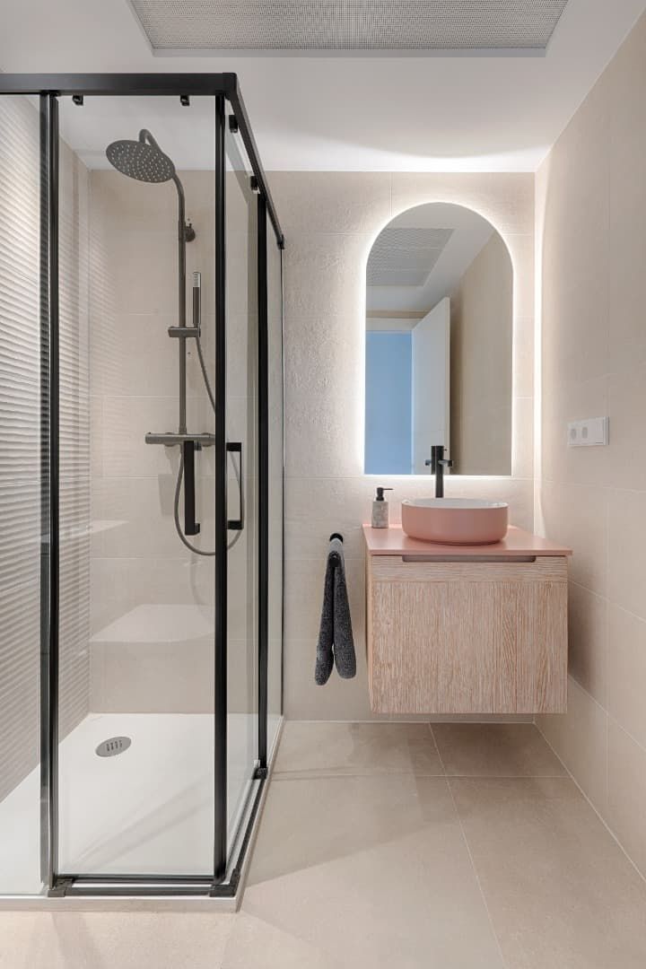 Baño moderno con ducha de cristal, lavabo flotante de madera, espejo redondo y suelo de baldosas beige.