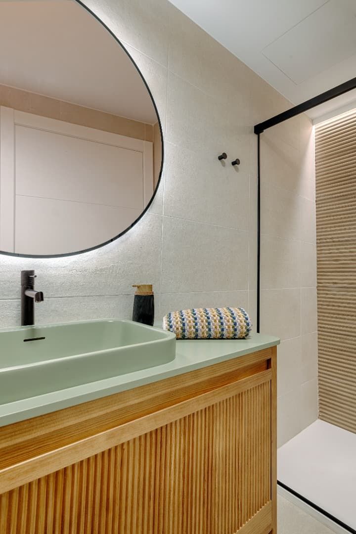 Tocador de baño moderno con espejo redondo, lavabo verde pálido y mueble de listones de madera bajo iluminación tenue.