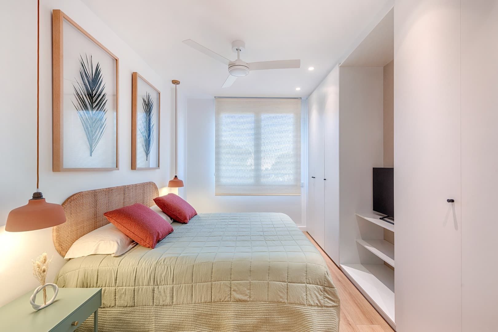 Habitación luminosa con cama, cuadro enmarcado, televisor empotrado y ventana con persianas blancas.