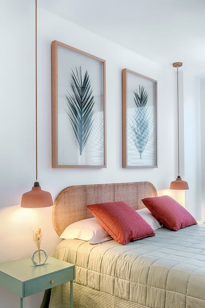Habitación acogedora con cama, almohadas rosas, lámparas colgantes de cobre y un cuadro de hojas enmarcado sobre una pared blanca.