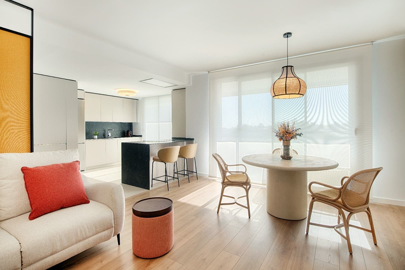 Cocina-comedor moderna y luminosa con muebles blancos, mesa redonda, sillas de mimbre y grandes ventanales.