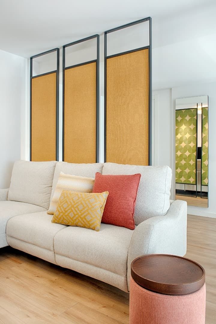 Sala de estar moderna con sofá color crema, cojines coloridos, suelo de madera y tres paneles altos enmarcados en la pared.